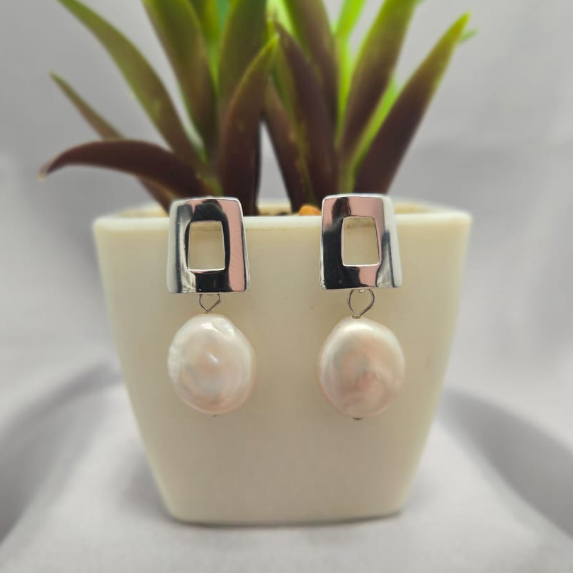Pearl earrings with silver hooks displayed in front of a small potted plant on a gray background