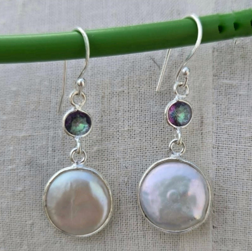 Pair of silver earrings with round gemstones hanging on a green stick against a textured fabric background.