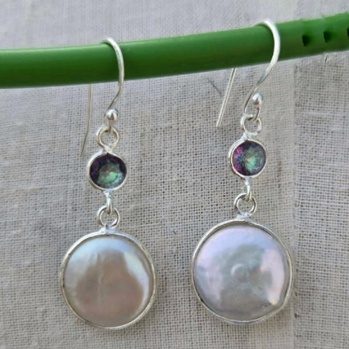Pair of silver earrings with round gemstones hanging on a green stick against a textured fabric background.