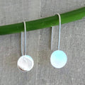 Silver earrings with round light blue stones hanging from a green rod against a textured gray background.