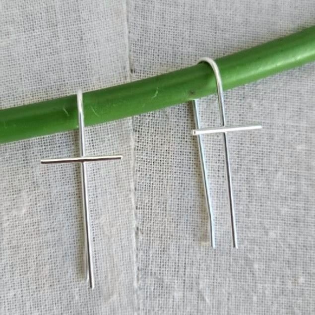 Silver cross-shaped earrings hanging from a green rod on a textured gray background