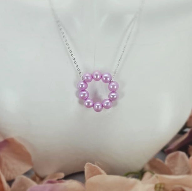 A sterling silver necklace with a pink freshwater pearl circle pendant, displayed on a white stand with pink flowers around it.
