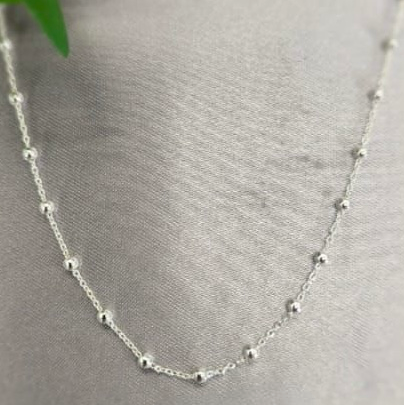 A silver ball chain necklace laid out on a textured surface with a green leaf in the background.
