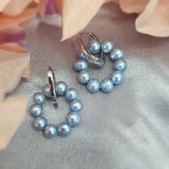 A pair of sterling silver huggie earrings attached to a blue freshwater pearl circle, displayed on a fabric surface with peach tones.