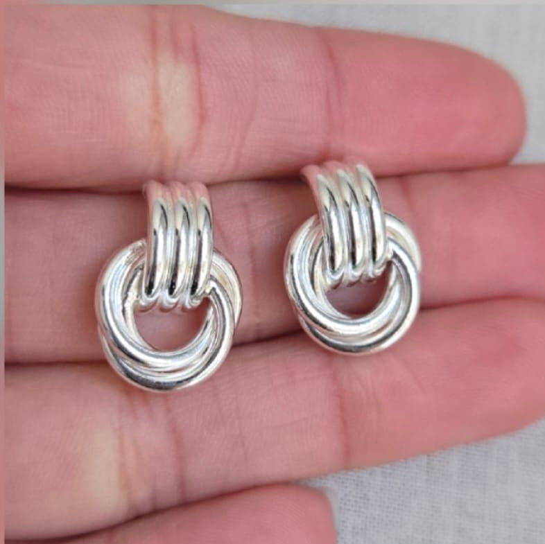 Silver knot earrings held between fingers on a neutral background