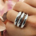 A person's hand wearing a bulky sterling silver three band ring with a glossy finish, set against a background of pink flowers.