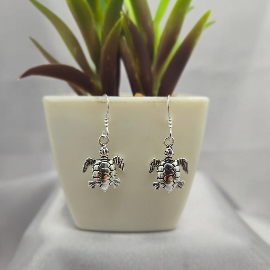 Silver turtle earrings on a light gray surface with a small plant in the background.