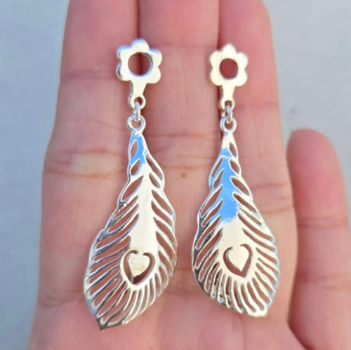Silver feather-shaped earrings with heart details held in a hand against a neutral background