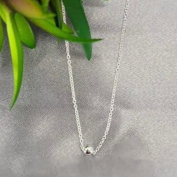 A sterling silver necklace with a tiny ball pendant, displayed on a grey background with a green plant to the side.