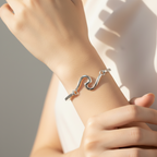 A sterling silver wave bracelet displayed on a white wristband, attached to a small artificial plant in a white vase with pebbles.