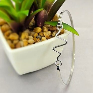 A sterling silver bracelet with a heartbeat design, displayed on a white surface next to a potted plant.