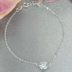 A sterling silver bracelet with a single cubic zirconia centerpiece, displayed on a grey surface with pink flowers in the background.
