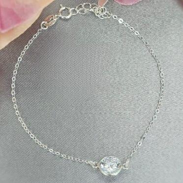 A sterling silver bracelet with a single cubic zirconia centerpiece, displayed on a grey surface with pink flowers in the background.
