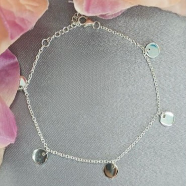A silver bracelet with five discs, featuring different embellishments on each disc, displayed on a surface with pink flower accents.