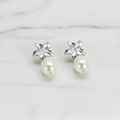A pair of sterling silver drop earrings with a frangipani flower design and freshwater pearl drops, displayed in a white vase with green leaves.