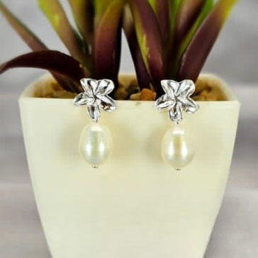 A pair of sterling silver drop earrings with a frangipani flower design and freshwater pearl drops, displayed in a white vase with green leaves.