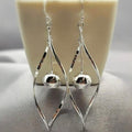A pair of silver drop earrings with a twist design and a dangling ball, displayed against a white background with a plant.
