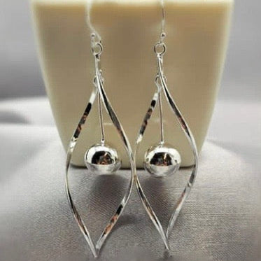 A pair of silver drop earrings with a twist design and a dangling ball, displayed against a white background with a plant.