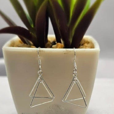 A pair of silver triangle-shaped drop earrings displayed on a white surface next to a potted plant.