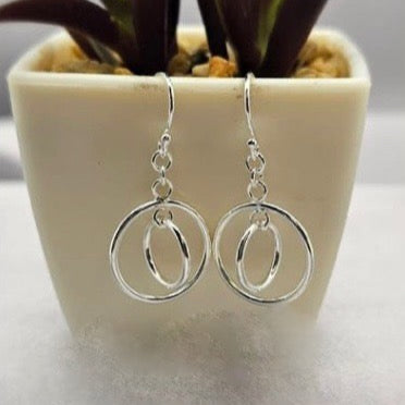 A pair of sterling silver dangling double circle drop earrings displayed in a white container with a green plant background.