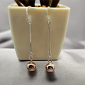 A pair of sterling silver chain earrings with rose gold balls on the ends, displayed on a table next to a white vase with a plant.