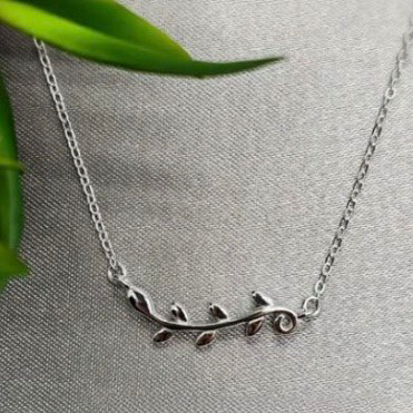 A sterling silver horizontal bar necklace with leaf patterns displayed on a textured surface with a green leafy background.