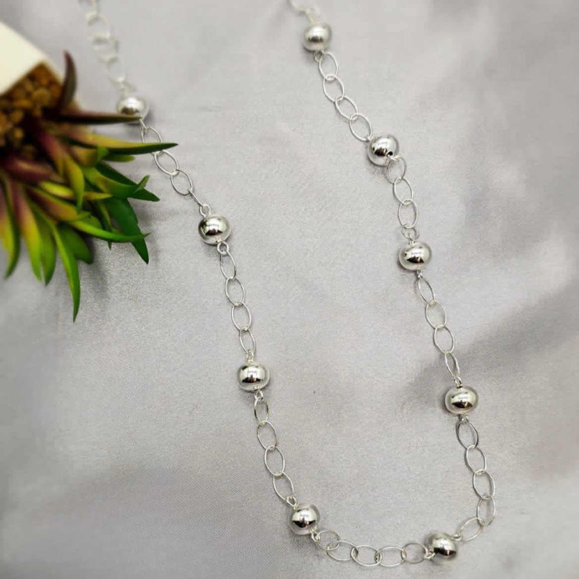 Silver beaded necklace on a gray surface with a plant in the background