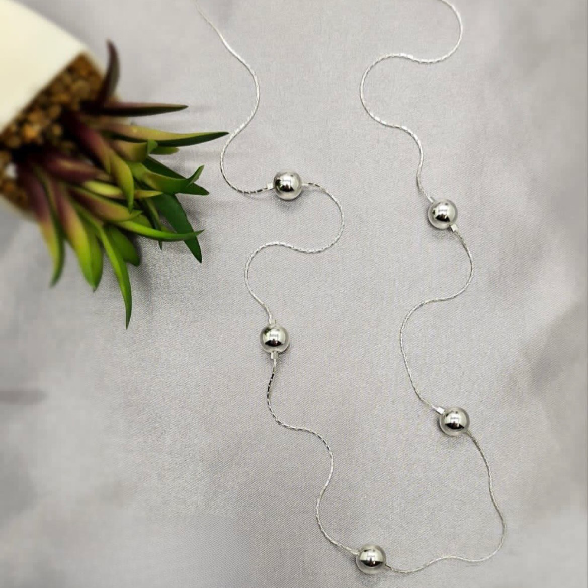 A sterling silver necklace with spherical pendants laid out on a flat surface with a floral patterned object to the side.