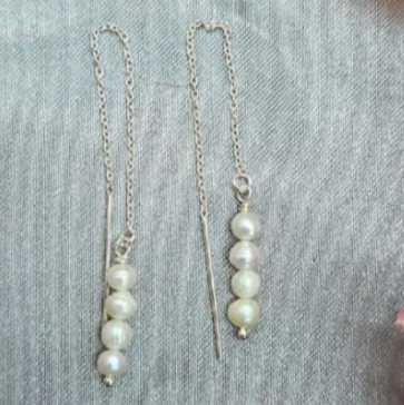 A pair of sterling silver thread earrings with four freshwater pearls on end, displayed against a neutral-toned textured background.