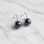 A pair of sterling silver hoop earrings with black freshwater pearl attachments displayed on a textured surface with a blurred flower in the background.