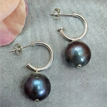 A pair of sterling silver hoop earrings with black freshwater pearl attachments displayed on a textured surface with a blurred flower in the background.