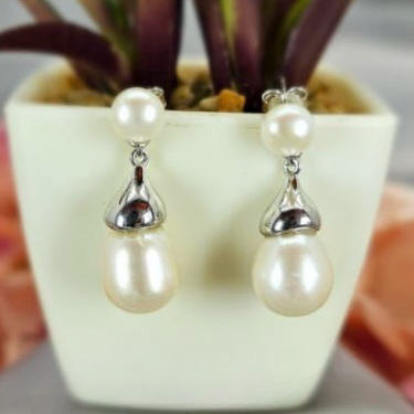 A pair of pearl earrings with silver hooks, displayed in a white vase with a green plant in the background.