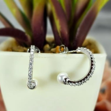 A pair of sterling silver hoop earrings with cubic zirconia pavé and freshwater pearls on end, displayed in a white vase with a green plant in the background.