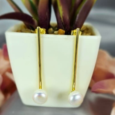 A pair of gold-plated on sterling silver earrings with freshwater pearls, displayed in a white ceramic vase with green plant leaves.