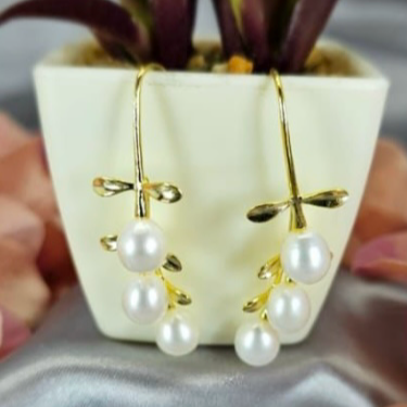 A pair of gold-colored drop earrings with pearl drops displayed in a white vase, with a green plant in the background.