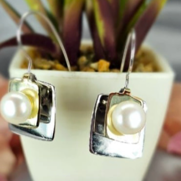 A pair of sterling silver statement earrings with gold accents and pearl detail, displayed in front of a potted plant.