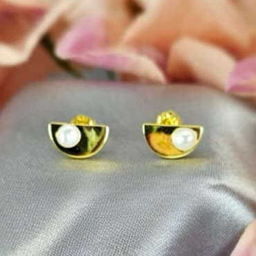 A pair of gold half circle stud earrings with freshwater pearls, placed on a silver surface with pink flower petals around.