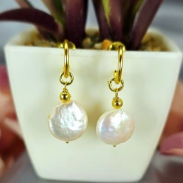 A pair of gold-plated on sterling silver earrings with pearl attachments, displayed in a white container with a green plant in the background.