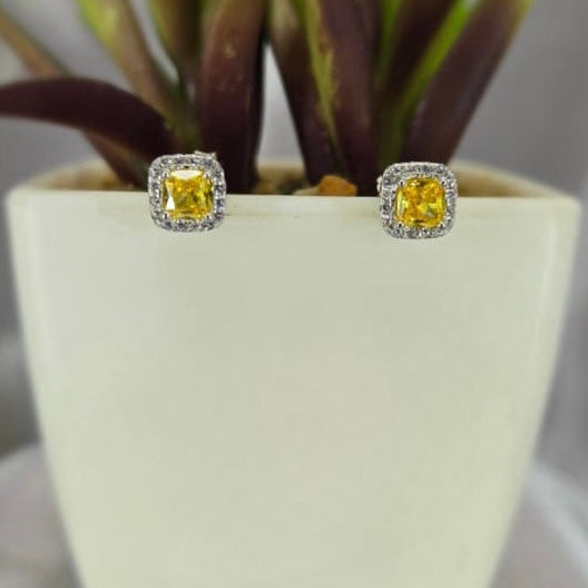 A pair of yellow square stud earrings with cubic zirconia, displayed in a white pot with a plant in the background.