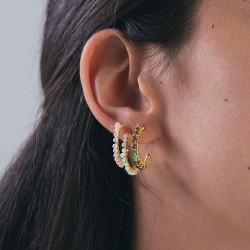 Close-up of an ear wearing gold hoop earrings with green gemstones against a neutral background