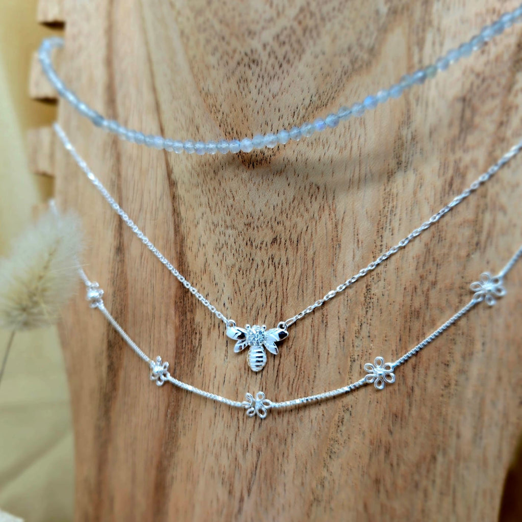 Two silver necklaces with bee and flower charms on a wooden surface