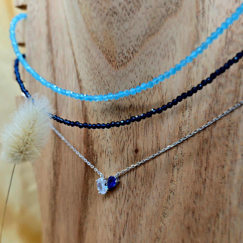 Three necklaces on a wooden surface, including a blue beaded necklace, a black beaded necklace, and a silver necklace with a blue pendant.