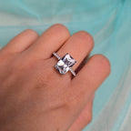 Hand wearing a diamond ring with a clear gemstone against a light blue fabric background