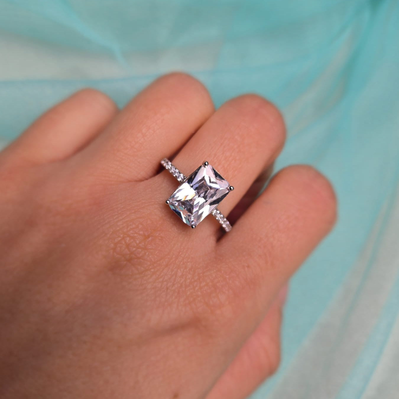 Hand wearing a diamond ring with a clear gemstone against a light blue fabric background