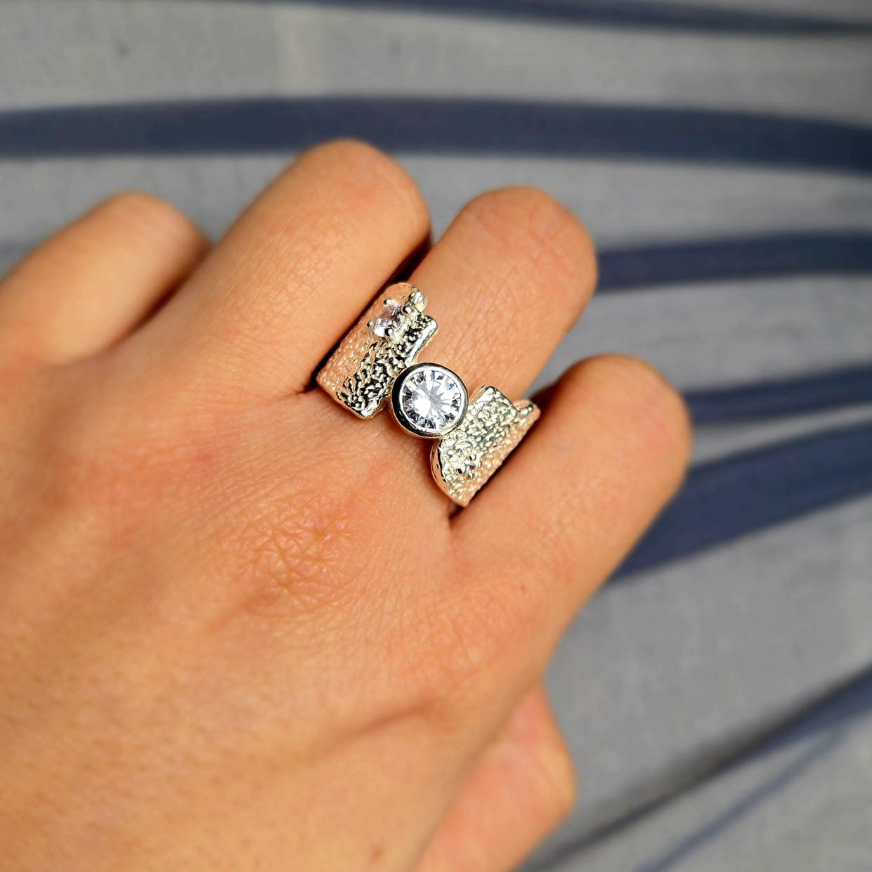 Close-up of a hand wearing two silver rings with a clear gemstone on a striped background