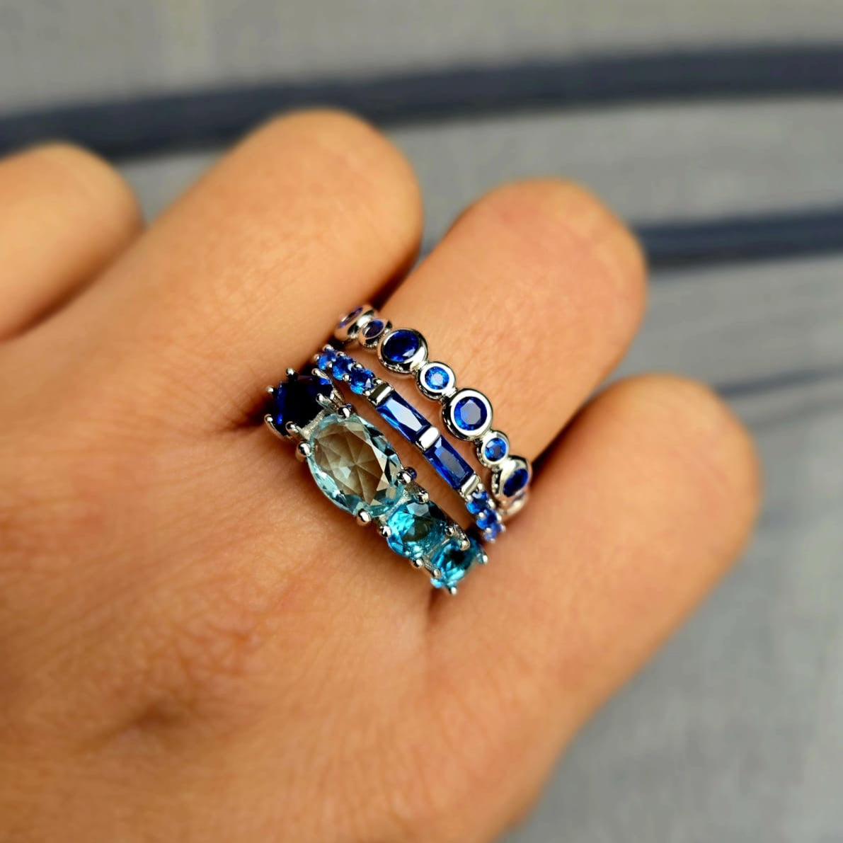 Close-up of a hand wearing a stack of blue gemstone rings on a neutral background