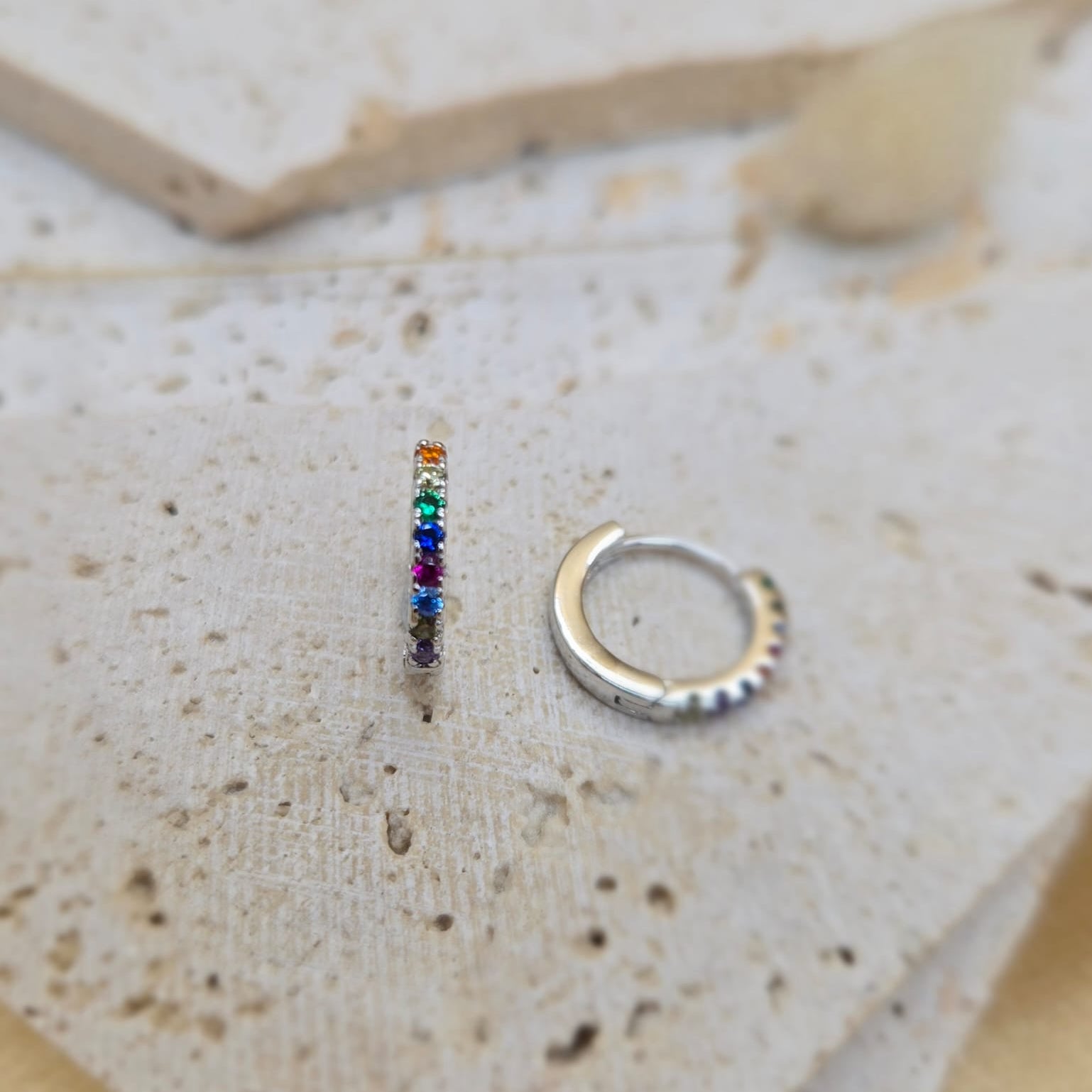 Small hoop earrings with colorful gemstones and silver finish on a textured surface