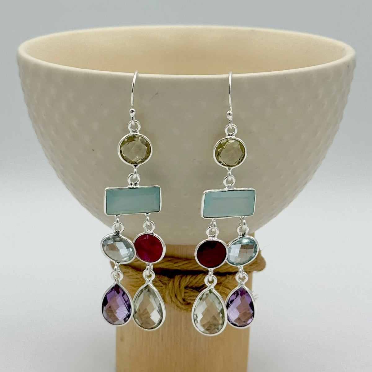 Multicolored gemstone earrings on a wooden stand with a ceramic bowl in the background
