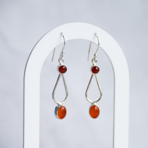 A pair of silver dangle earrings with small round red beads, elongated silver teardrop shapes, and oval orange-red stone drops, displayed on a white arched stand against a light gray background.