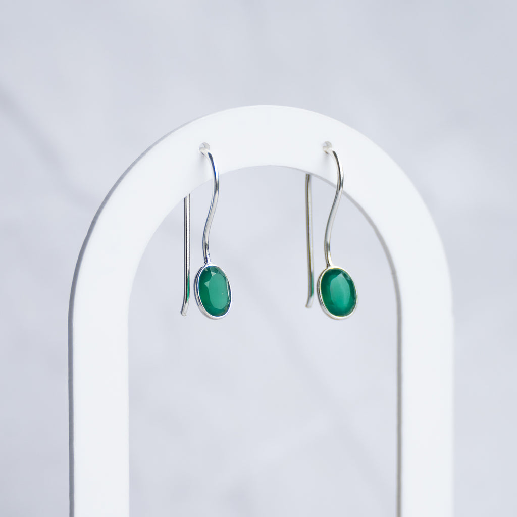 A pair of silver drop earrings with oval green cabochon stones, hanging from a white arched jewelry display stand against a light gray marble background.
